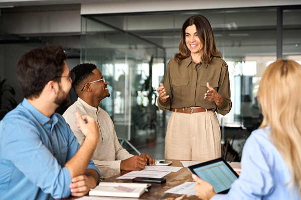 Besprechungssituation in einem Büro – eine Frau spricht und sieht zu 3 sitzenden Personen – Marktforschungsmethoden in Online-Panels