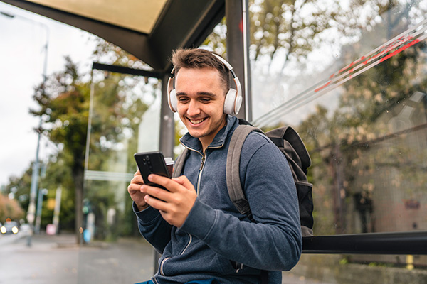 Mann mit Kopfhörern hält Smartphone – sitzt auf Bushaltestelle